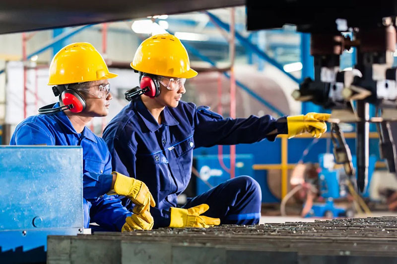 Construction worker wearing a safety helmet, work uniform, earmuffs, goggles, and gloves on-site.