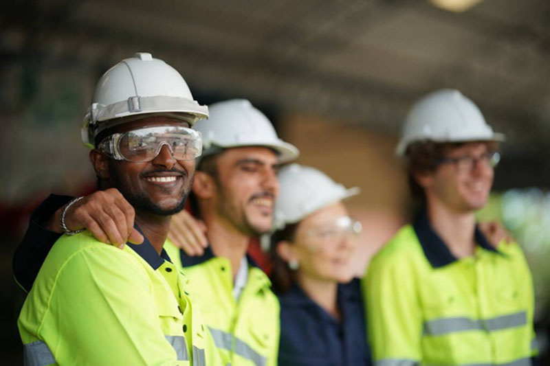 Group of smiling employees in reflective long-sleeve gear, safety helmets, and glasses, promoting ergonomic and comfortable PPE.