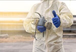 A worker in disposable isolation gown, holding a respirator mask and wearing gloves, giving a thumbs up to demonstrate confidence in PPE reliability.