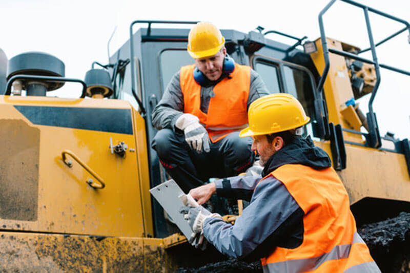 Two workers wearing PPE (protective clothing, reflective vests, helmets, and ear protection) discussing safety measures in a high-risk work environment.