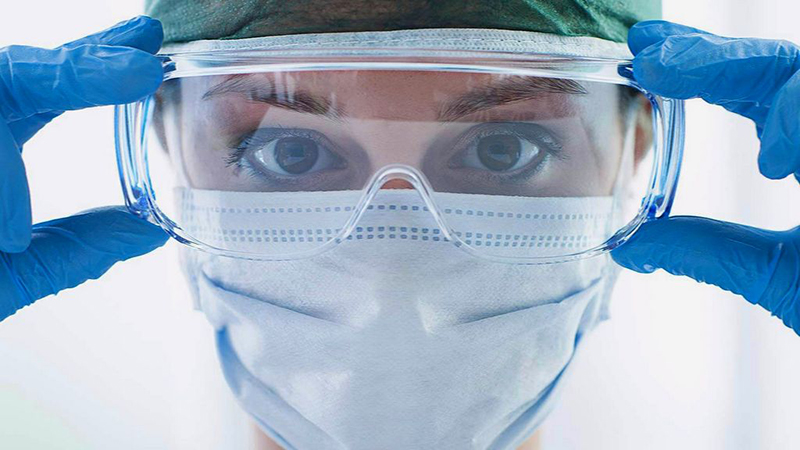 Healthcare worker adjusting protective goggles while wearing medical PPE, including a face mask and gloves.